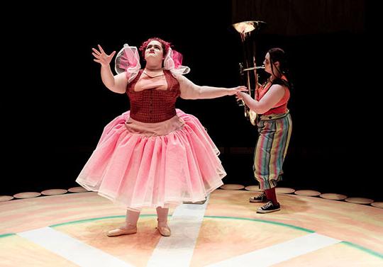 woman on stage acting, someone playing tuba behind them